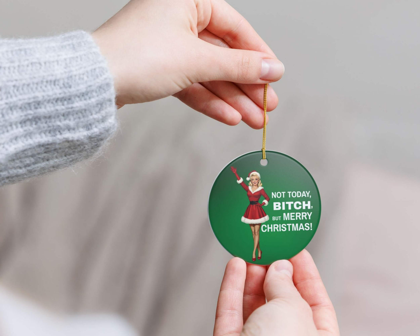 Green Christmas ornament with a humorous design and text, held by two hands against a neutral background.